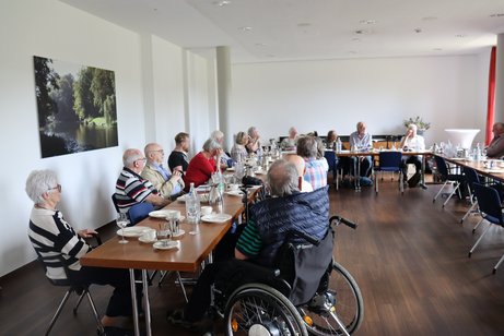 Mehrere Personen sitzen an langen Tischen und h&ouml;ren dem Vorsitzenden zu.