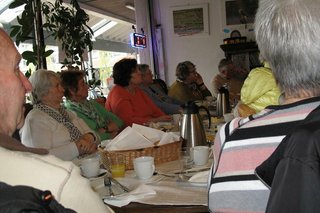 Viele Menschen sitzen gemeinsam in einem Raum an einem Tisch und hören einem Vortrag zu.