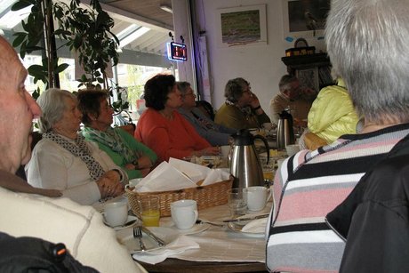 Viele Menschen sitzen gemeinsam in einem Raum an einem Tisch und hören einem Vortrag zu.