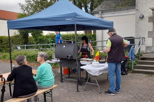 Auf dem Bild sind ein Grill mit Grillgut vor einem Pavillon zu sehen. Einige Leute stehen drumherum.