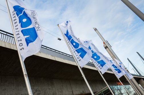 Vier VdK-Flaggen wehen an Masten im Wind, im Hintergrund Himmel und ein Teil einer Brücke. 