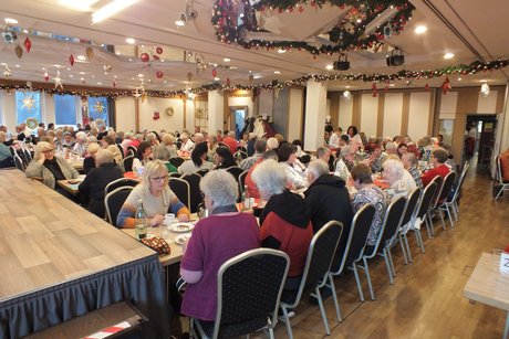 In einem gro&szlig;en Raum sitzen Menschen an langen, festlich gedeckten Tischen.