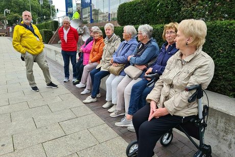 Eine Gruppe Menschen sitzt nebeneinander auf einer langen Bank vor einer Kirschlorbeerhecke.