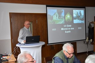 Konrad Eilers steht am Rednerpult neben der Leinwand und berichtet mit Beamer-Unterstützung über die Historie des Westumer Kapellchens.