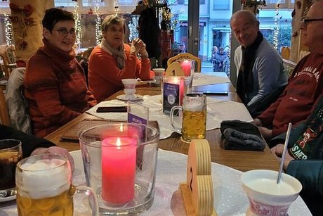 Eine Gruppe Männer und Frauen sitzt in einer Gaststube bei Heiß- und Kaltgetränken an einem langen Tisch.