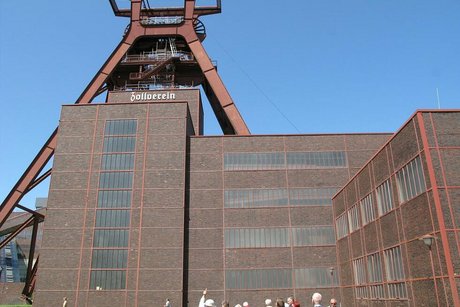 Eine Gruppe Menschen steht klein im Bildvordergrund vor dem markanten Zechengebäude mit Förderturm und der weißen Aufschrift Zollverein
