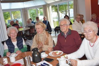 Mitglieder sitzen am Tisch und trinken Kaffee und essen Kuchen. Dabei hören sie aufmerksam zu. Auch der Ehrenvorsitzende Manfred Radermacher sitzt am Tisch.