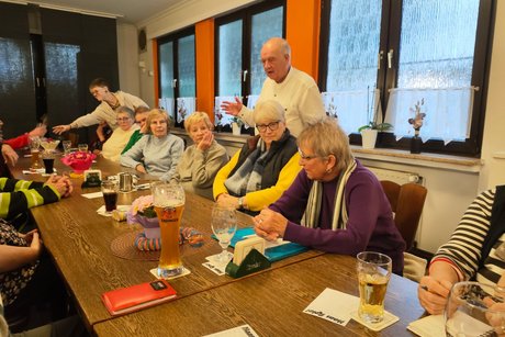Mehrere Menschen sitzen an langen Tischen und h&ouml;ren einem Mann zu, der einen Vortrag h&auml;lt.