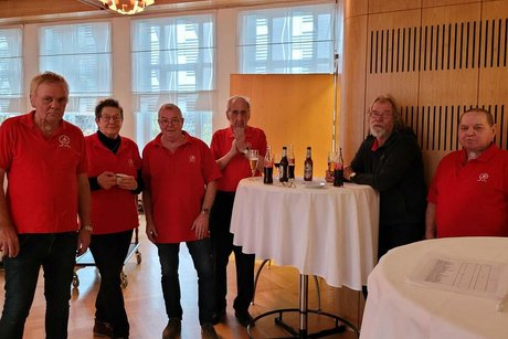 Sechs Männer des Chors "Die Kehlkopflosen" in roten T-shirts an einem Stehtisch.
