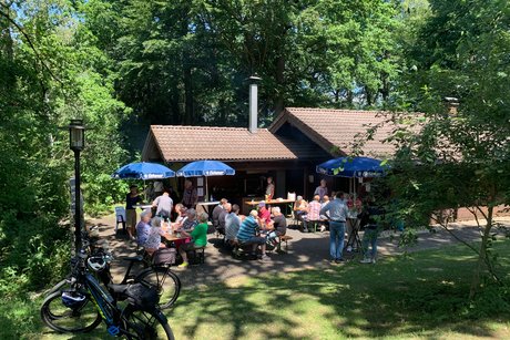 Grillh&uuml;tte im Gr&uuml;nen mit Personen an Tischen unter Sonnenschr&iacute;rmen mit Fahrr&auml;dern im Vordergrund