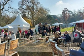 Bierzeltgarnituren und Holztische und Stühle sind auf einem Platz aufgestellt. Rundherum flanieren Menschen entlang von Weihnachtsmarktbuden und weißen Pavillon-Zelten.