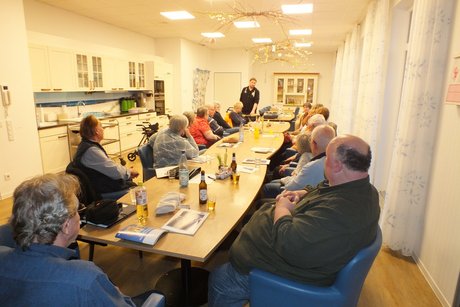 Ein Mann steht vor einem langen Tisch , an dem viele Menschen sitzen.
