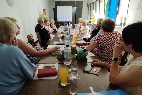 Verkehrsquiz mit Polizeihauptkommissar Hans-Joachim Ruhl. Mehrere Personen sitzen an einem langen Tisch und hören Polizeihauptkommissar Hans-Joachim Ruhl zu.