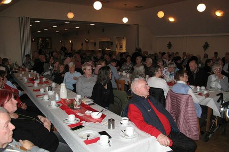 Viele Menschen sitzen in einem Saal an langen, weihnachtlich geschmückten Tischen.