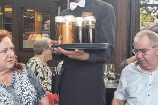 Ein junger Ober präsentiert lächelnd ein Tablett mit frischen Bieren in Richtung Kamera. Rechts und links von ihm sitzen eine Frau und ein Mann.