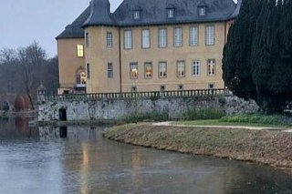 Eine leichte Eisdecke überzieht den Wassergraben des Schlosses, dessen Fenster im Bildhintergrund in der Dämmerung leuchten.