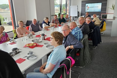 20 Teilnehmer sitzen an einer Kaffeetafel und der Mitarbeiter erklärt per Beamer den Wertegang der Firma