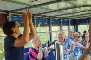 Eine Frau steht im Planwagen und hat die Hände zum Klatschen erhoben. Im Hintergrund sitzen weitere Menschen und klatschen.