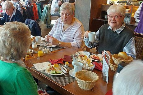 Zwei Frauen sitzen gemeinsam beim Frühstück an einem Tisch. Ihnen gegenüber sitzen weitere Menschen und im Hintergrund erkennt man weitere Tische mit Gästen des Cafés.