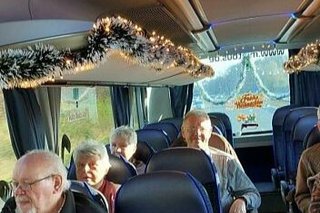 Menschen sitzen in einem Reisebus, dessen Gepäckablage mit Lichterketten und Weihnachtsgirlanden geschmückt ist.