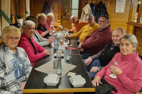 Eine Gruppe Männer und Frauen sitzt in einer Gaststube an einem langen Tisch beisammen.