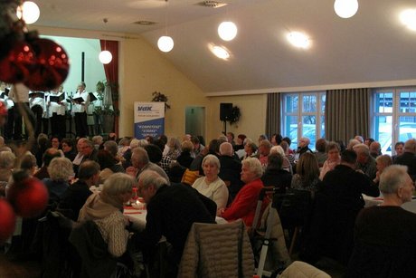 Viele Menschen sitzen in einem Saal an langen, weihnachtlich geschmückten Tischen. Links im Bild schimmern rote Weihnachtskugeln
