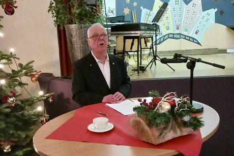 Ein Mann steht an einem großen runden Stehtisch, auf dem sich ein Adventsgesteck befindet. Im Hintergrund links erkennt man die Zweige eines geschmückten Weihnachtsbaumes, im Hintergrund befindet sich eine Bühne.