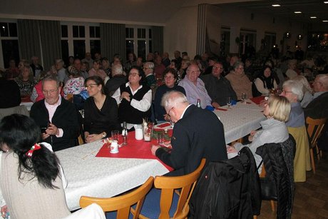Viele Menschen sitzen gemeinsam in einem Saal an weihnachtlich geschmückten, langen Tischen.