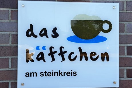 Ein wei&szlig;es Schild auf einer Mauer mit schwarzer Schrift das K&auml;ffchen am Steinkreis