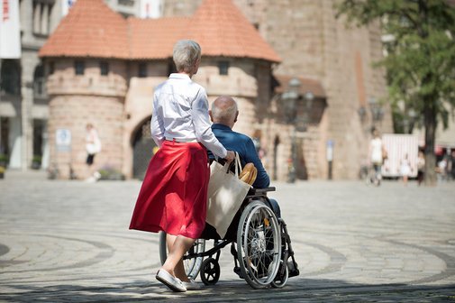 Paar in der Stadt unterwegs Eine Seniorin schiebt ihren Mann, der im Rollstuhl sitzt, über einen Platz, man sieht im Hintergrund ein altes Stadttor.