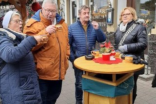 Zwei Männer und zwei Frauen stehen an einem Stehtisch aus Holz. Der eine Mann raucht gerade eine Zigarette. Die Frau und der Mann links im Bild prosten sich mit Glühweintassen zu.