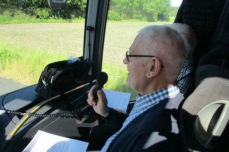 Ein Mann sitzt vorne als Beifahrer im Reisebus und spricht in das Bus-Mikorfon.