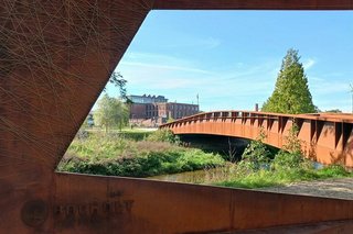 Ein rostiger Stahlrahmen rahmt den Blick auf einen Kanal, über den sich eine rostige Stahlbrücke spannt.