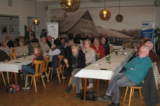 Menschen sitzen in einem Saal an langen Tischen und hören einem Vortrag zu.