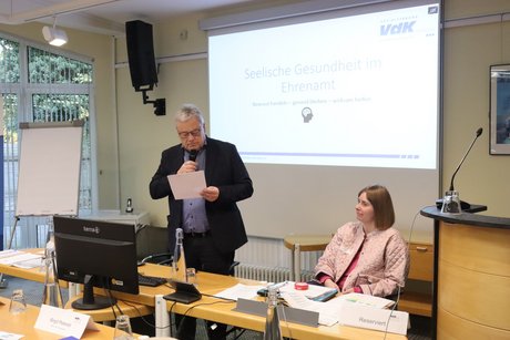 Ein Mann spricht und eine Frau sitzt daneben.