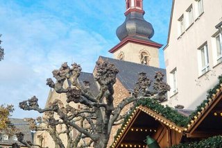 Der Kirchturm von Rüdesheim hebt sich vor blauem Himmel ab. Entlang der Häuserfassade und zwischen zurückgeschnittenen Platanen reihen sich Weihnachtsmarktbuden aneinandner.