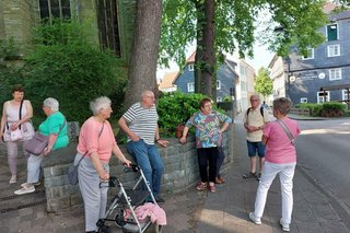 Eine Gruppe Menschen steht, lehnt oder sitzt an, bzw. auf einer Mauer und wartet.