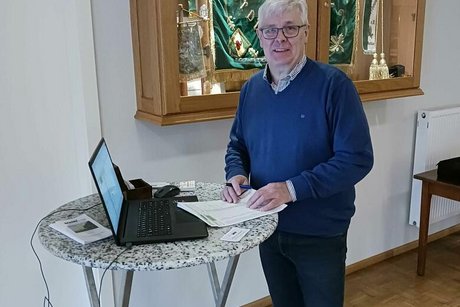 Ein Mann steht an einem Stehtisch auf dem ein Laptop und Papiere liegen. Im Hintergrund befindet sich ein beleuchteter Schaukasten mit Stoffbannern.