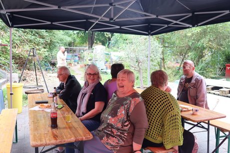 Mitglieder und Gäste im Außenbereich. Mehrere Menschen sitzen im Außenbereich an Tischen und unterhalten sich angeregt.