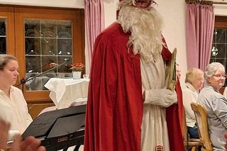 Im Vordergrund ein rot gekleideter Nikolaus mit Mitra auf de Kopf, im Hintergrund eine Frau am Keyboard