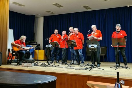 Sechs Männer des Chors "Die Kehlkopflosen" stehen, begleitet von einem Gitarristen auf der Bühne und singen Lieder.