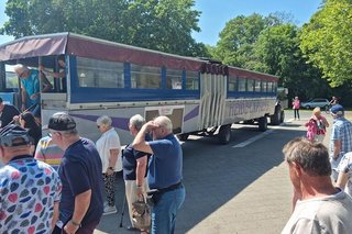 Menschen stehen auf einem gepflasterten Platz, im Hintergrund wartet ein großer, zweizügiger Planwagen auf Abfahrt.