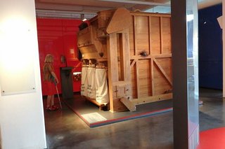 Eine raumgroße Apparatur aus Holz steht in einer Ecke der Ausstellung. An der Frontseite sind drei große Stoffbeutel befestigt.