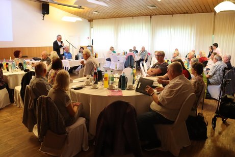 Viele Menschen sitzen an gro&szlig;en runden Tischen.
