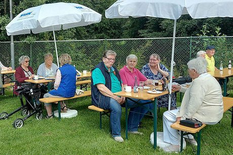 Menschen sitzen gemeinsam bei Getränken unter Sonnenschirmen an einer Bierzeltgarnitur.