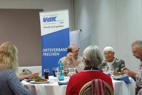 Männer und Frauen sitzen gemeinsam an einem Tisch. Auf dem Tisch stehen Getränke und Teller mit Grünkohl.