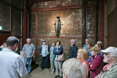 Eine Gruppe Menschen steht in einem Raum mit roten Ziegelwänden. An der Stirnseite des Raumes hängt, von Deckenstrahlern beleuchtet, eine Bronzefigur der heiligen Barbara, Schutzpatronin der Bergleute.