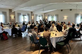 Menschen sitzen an Tischen in einem Saal. Am Rand steht ein Mann mit Mikrofon und spricht