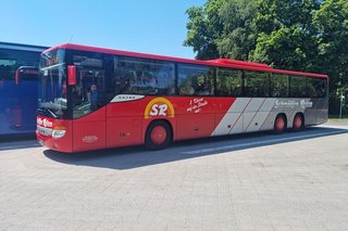 Ein großer roter Reisebus steht abfahrtbereit auf dem gepflasterten Platz vor dem Ressort.