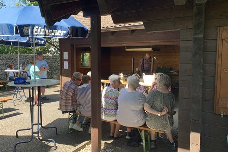 Personen an einem Tisch vor einer Grillh&uuml;tte unter Sonnenschirmen